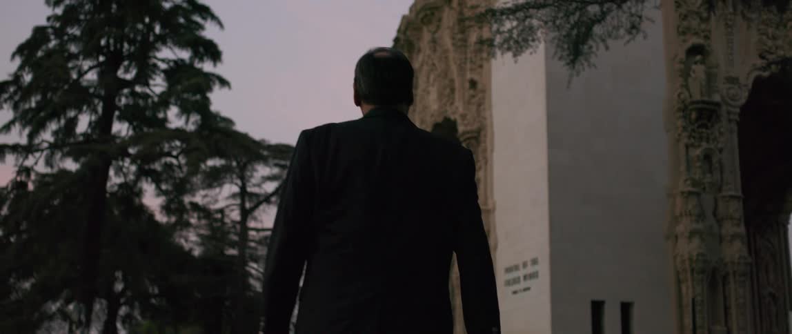 An elderly man in a dark suit is walking towards a monument in a park
