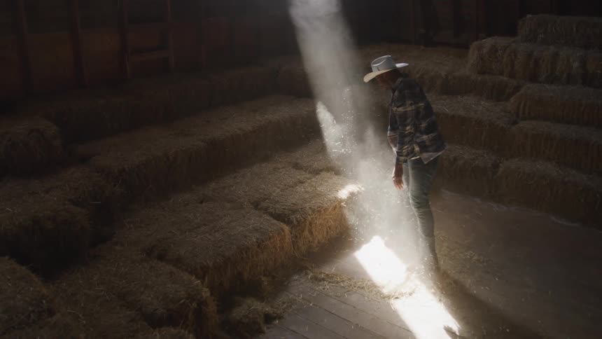 A farmer wearing a sunhat inside a storage unit tossing a bunch of straw beside other stored straw