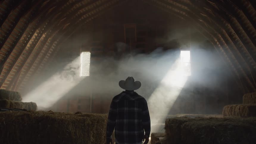 A person wearing a cowboy hat and a plaid shirt walks inside a dusty barn with hay bales illuminated by beams of light entering through openings in the roof