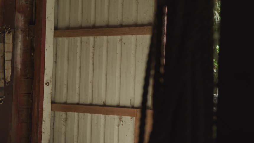A man in a cowboy hat and plaid shirt closes the door to a barn or stable