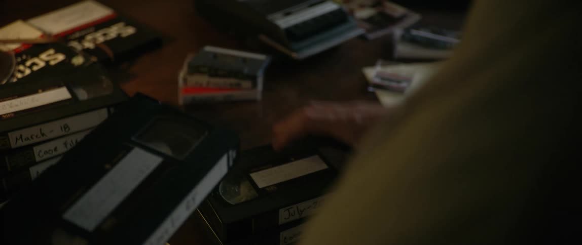 A man sitting behind a desk sorting through a series of vhs tapes and cassette tapes on the table