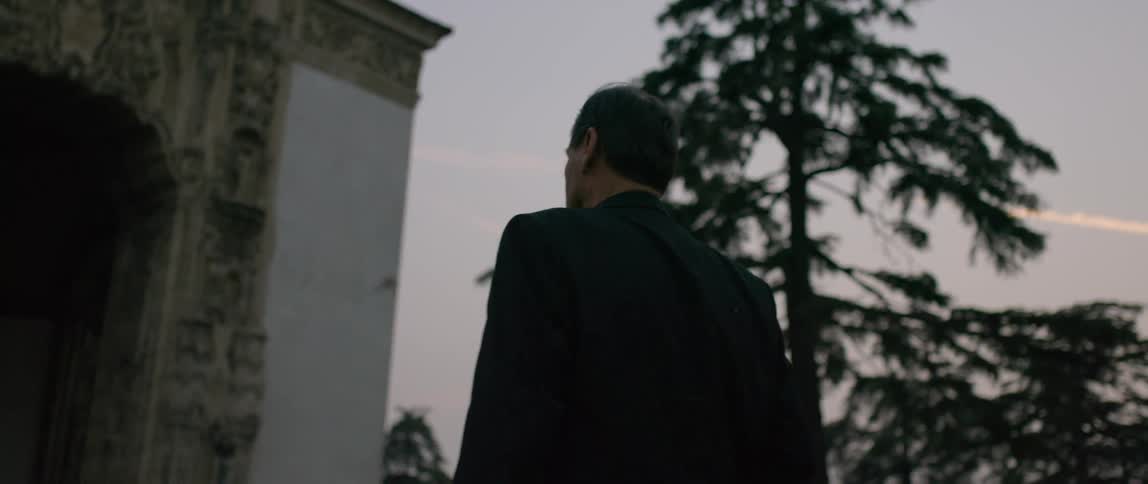 A man in a dark suit is walking slowly towards a mausoleum on a cloudy evening