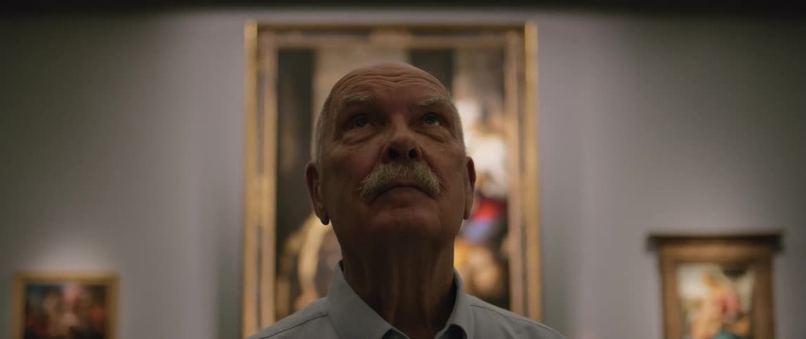 The face of an elderly man looking forward in an indoor setting with a huge painting behind him