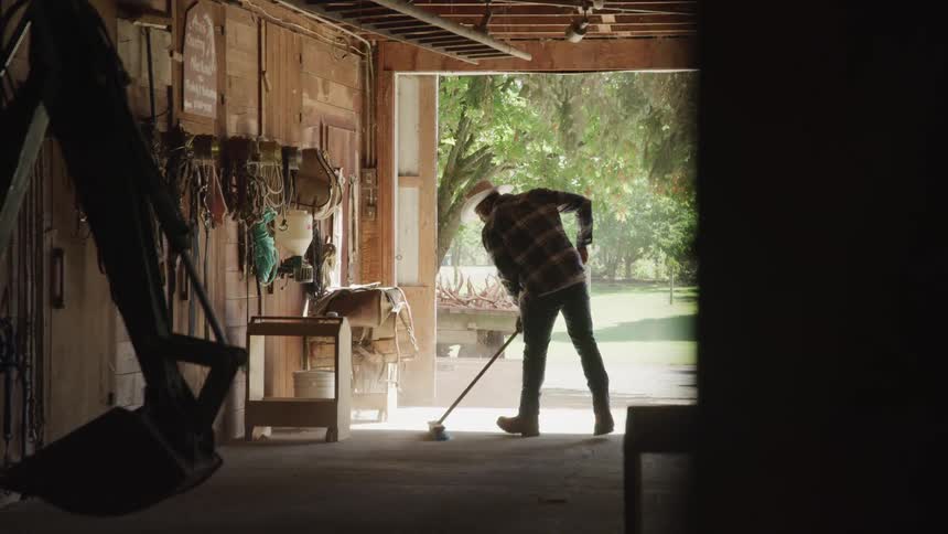 A man is using a push broom to sweep a workshop with construction equipment in it