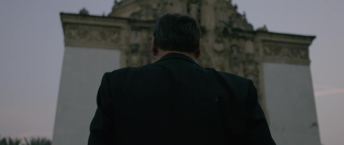 A man in a dark suit walks towards a large ornate building under a dusky sky