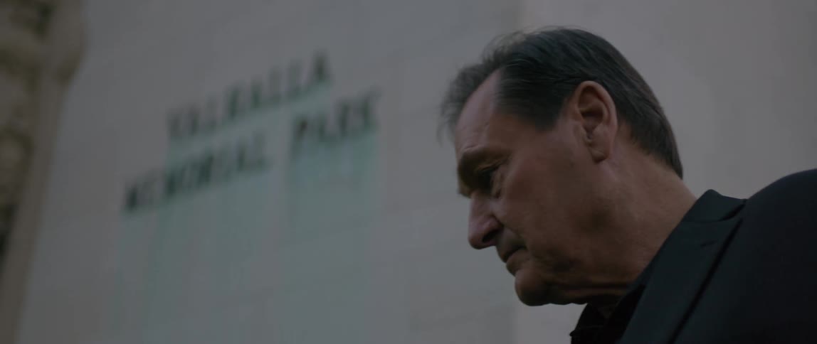 Closeup of a man standing near a white wall with a sign reading valhalla memorial park streaked with green stains running down its letters