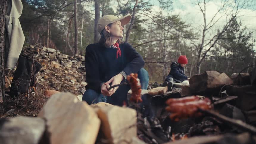 Woman cooking hot dogs over a campfire while camping in the woods with her son
