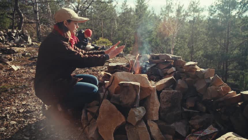 At the top of a forest a woman warms herself by a campfire while a boy in the background writes on a piece of paper
