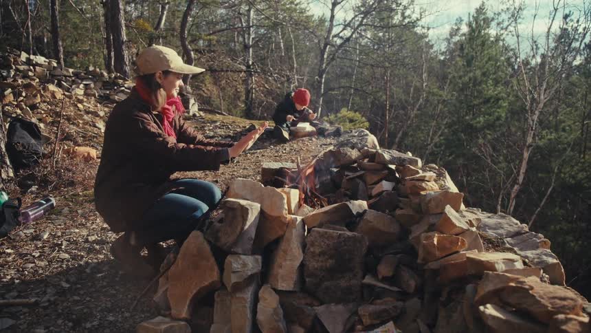 Woman warming her hands near a campfire in the woods as her son plays nearby