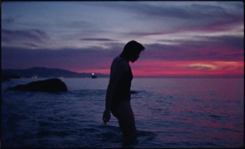 A silhouetted figure walks into the ocean as the sun sets casting a gradient of pinks and purples across the water and sky