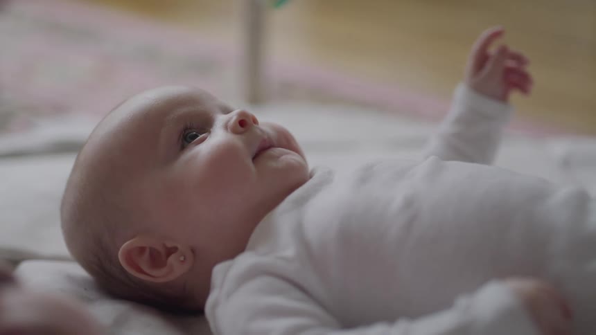 A baby wearing a white onesie lies on a patterned blanket with a small earring visible in one ear