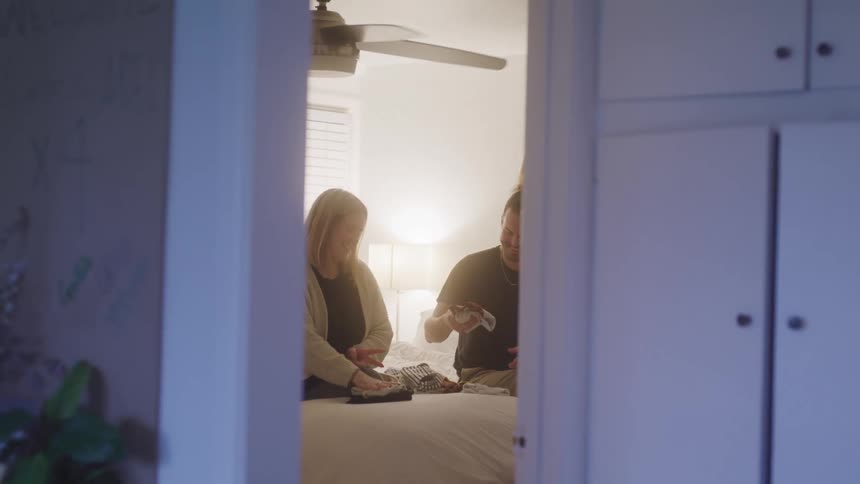 A couple sits together on a bed folding baby clothes