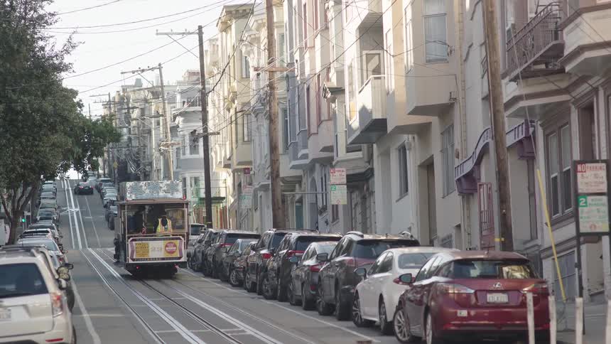 A trolley drives down a street lined with older houses and parked cars