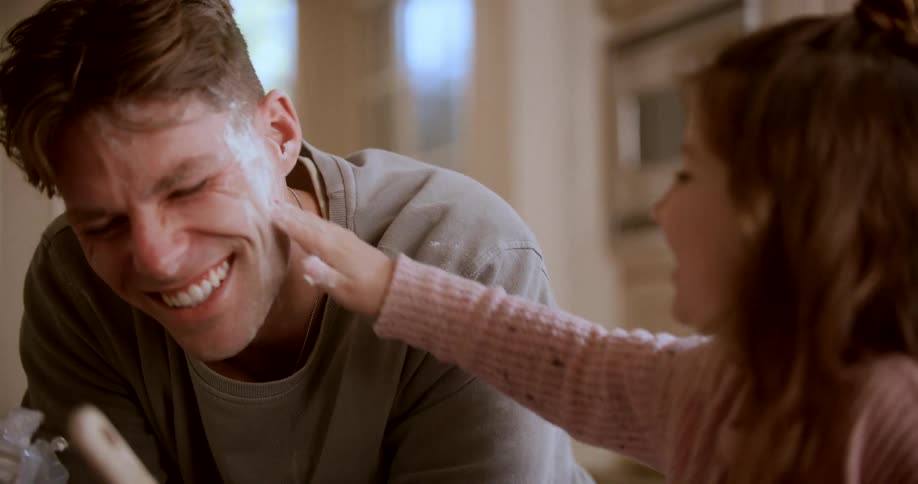 A young father in a sunny kitchen laughs as his small daughter playfully smears his face with flour