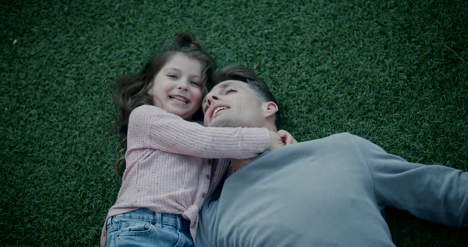 Happy little girl laughing as she hugs her dad while both laying on the grass outdoors