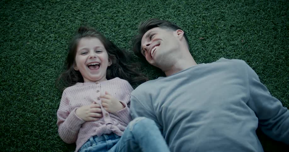 A young father and his daughter lie playfully next to each other on the grass both looking happy and carefree