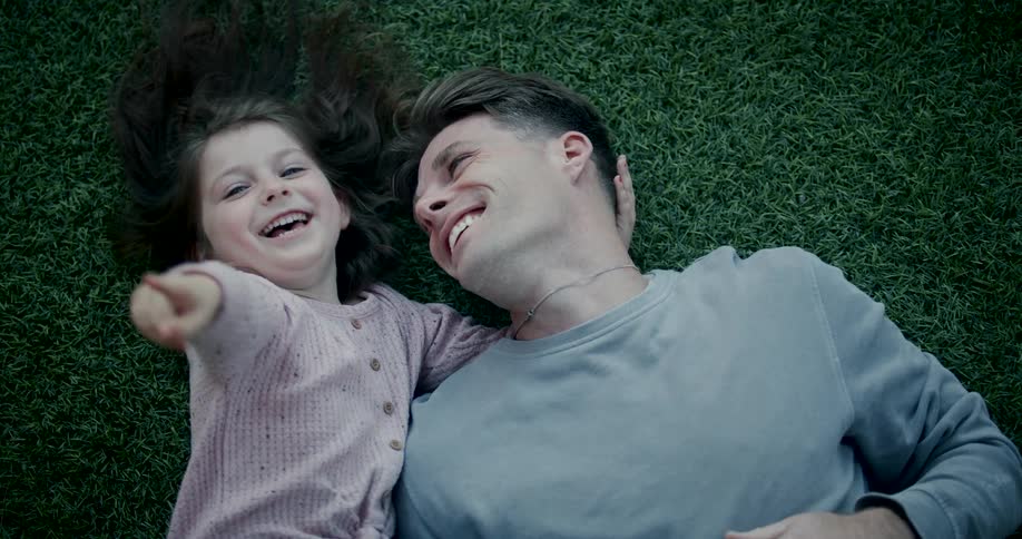 A young man and his daughter lie side by side on short grass both laughing as the daughter points to the sky while holding her father behind his head at midday