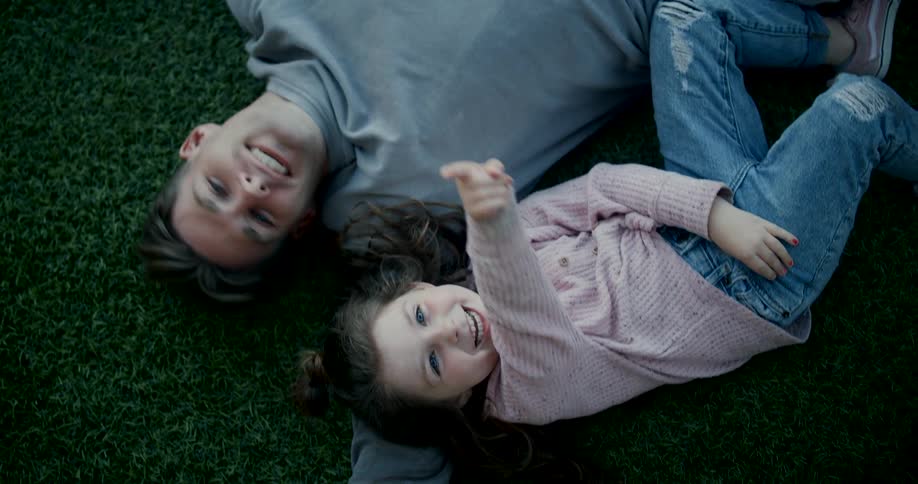 A young man and his daughter lie side by side on short grass at midday both laughing as the daughter points to the sky