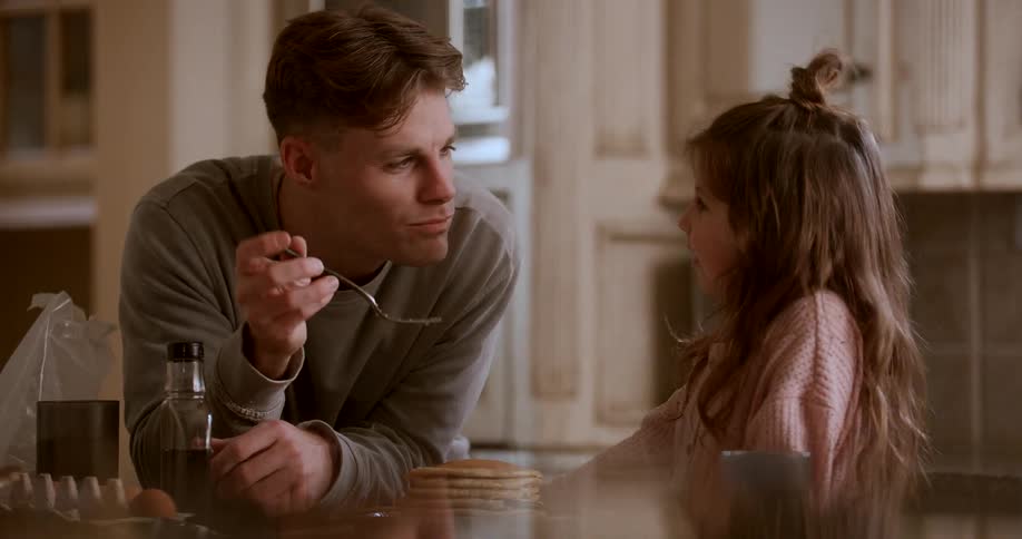 A father and daughter stand at the kitchen counter while the father eats pancakes