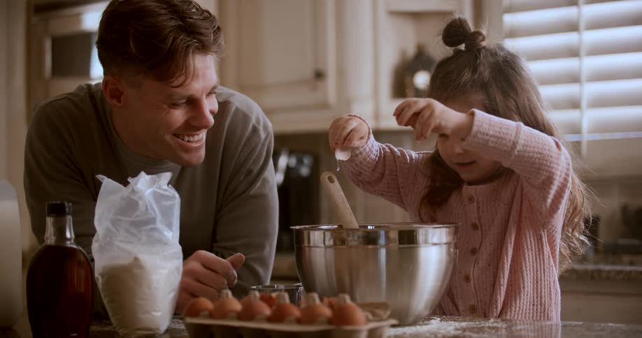 A father smiles while his young daughter cracks an egg into a mixing bowl