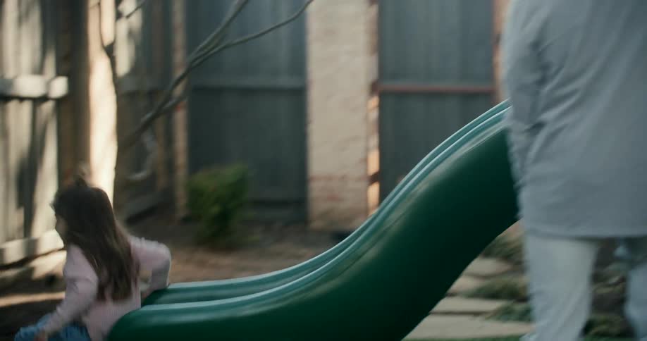 A father watches his young daughter slide down a toboggan in a backyard play area on a sunny morning