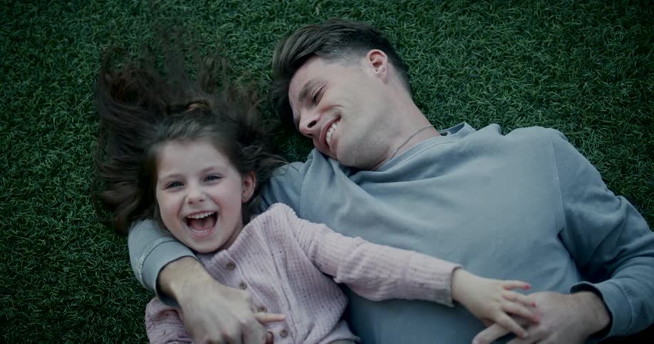 A young father and his daughter lie on the grass sharing a joyful and carefree moment together