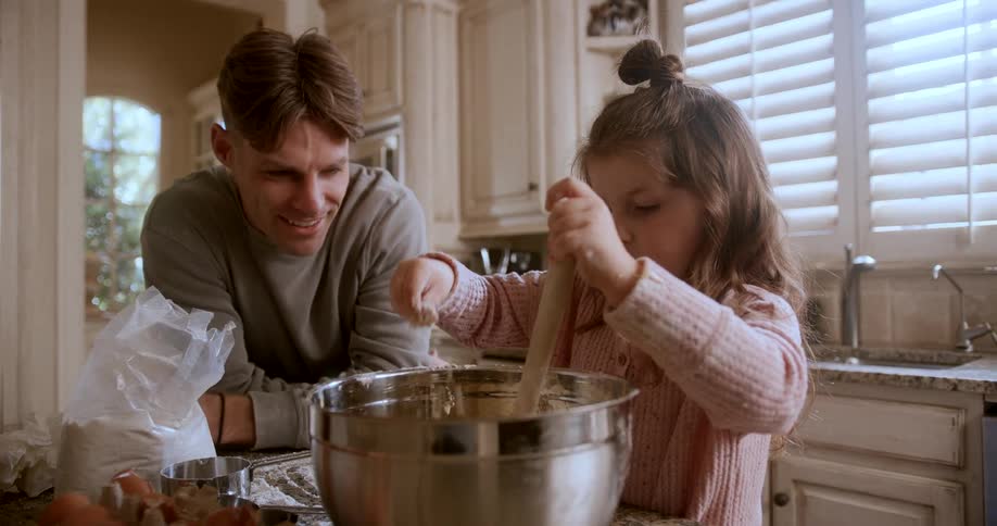A father helping and watching his daughter a child stirring contents in a bowl while in the kitchen at midday