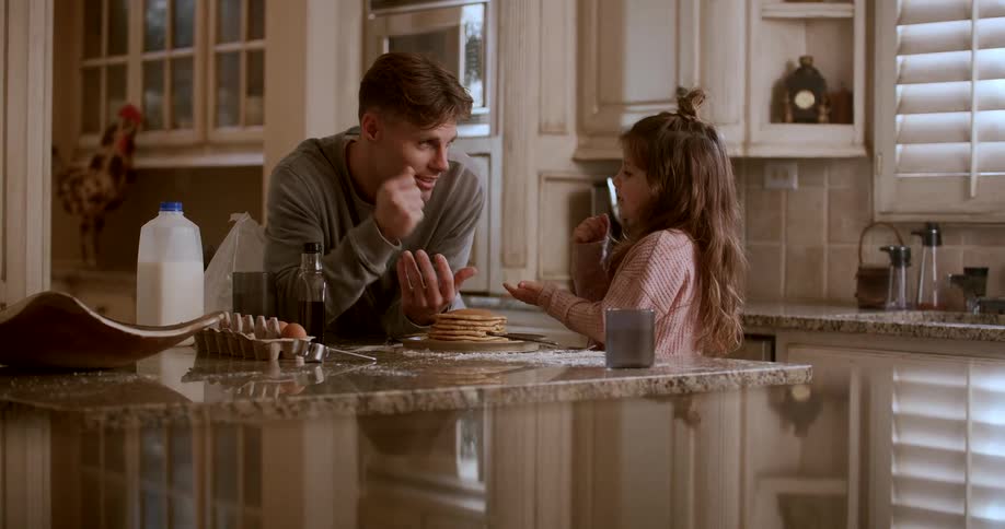 A father and daughter interact enthusiastically while standing at a kitchen counter with a stack of pancakes between them