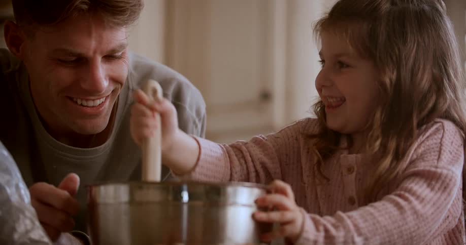A man happily watching his daughter mix contents in a bowl playfully