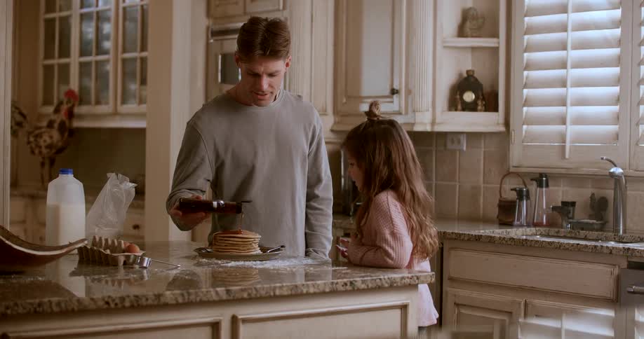 Dad pouring maple syrup over a stack of pancakes on kitchen counter while his daughter watches