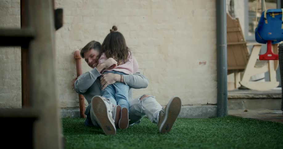 A father sits in the grass playing with his daughter as she jumps and topples him they fall together laughing happily in the backyard