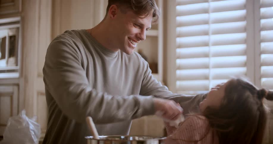 A father is cleaning his daughters face with a napkin as they both laugh together in the kitchen at midday