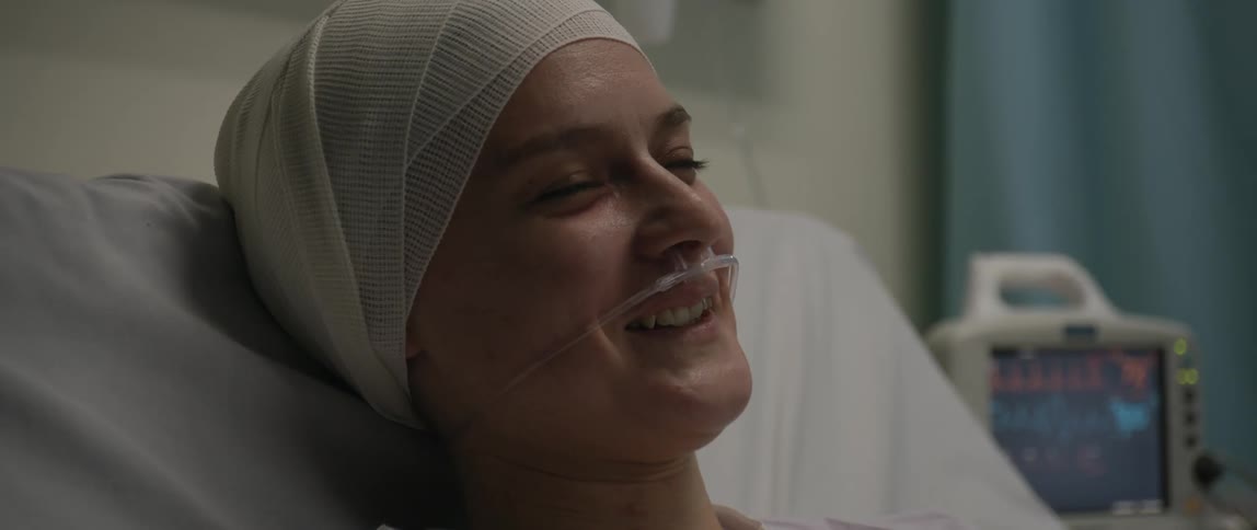 A woman lying on a hospital bed turns towards the camera and smiles
