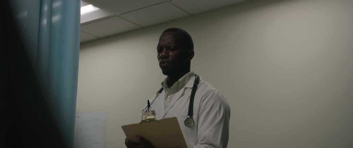 A doctor in a white coat and stethoscope stands in a room holding a clipboard then walks away