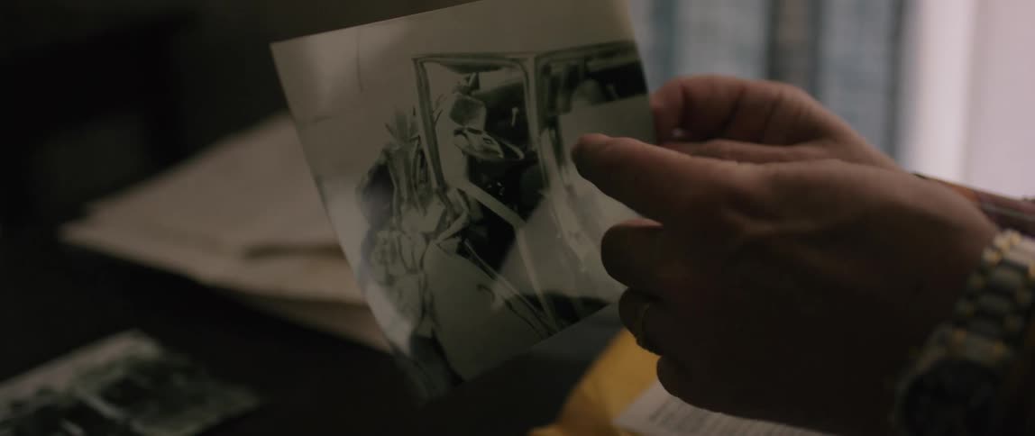 A hand wearing a watch and ring points at a blackandwhite photograph of a car accident on a table scattered with other photographs
