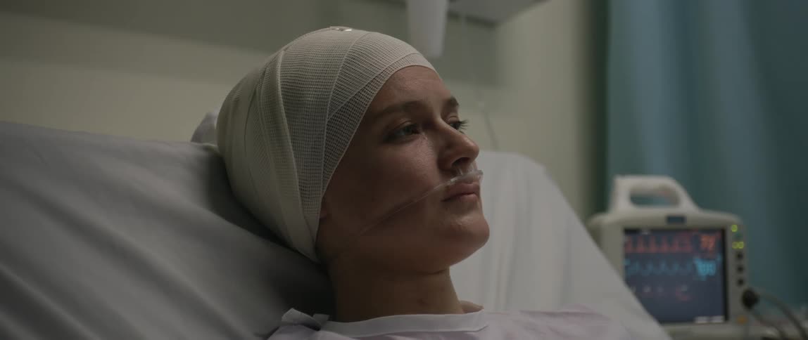 Closeup of a woman in a hospital bed with her head fully bandaged and a nasal cannula alongside a nearby heart rate monitor