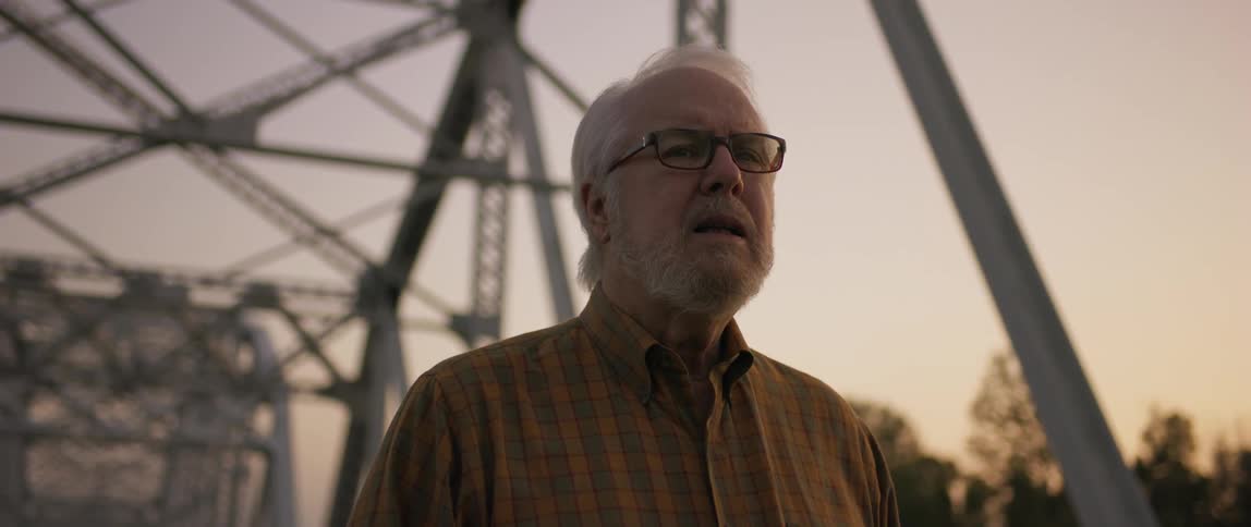 Elderly man is standing outdoors near a bridge at sunset