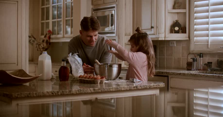 Father smiles while watching his daughter add flour to a metal bowl standing on their kitchen island