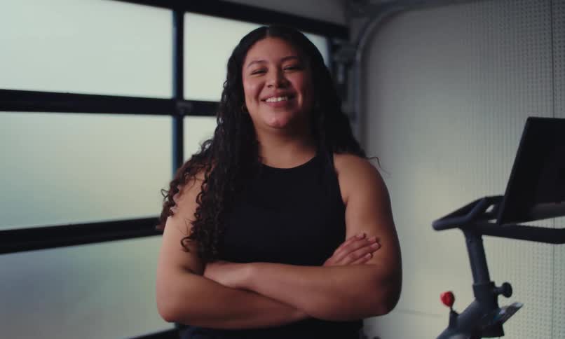 A hispanic woman with long hair is standing near a stationary bike in a garage