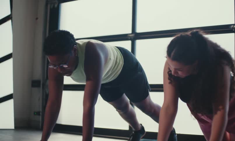 Two women working out with light hand weights while doing planks on a yoga mat