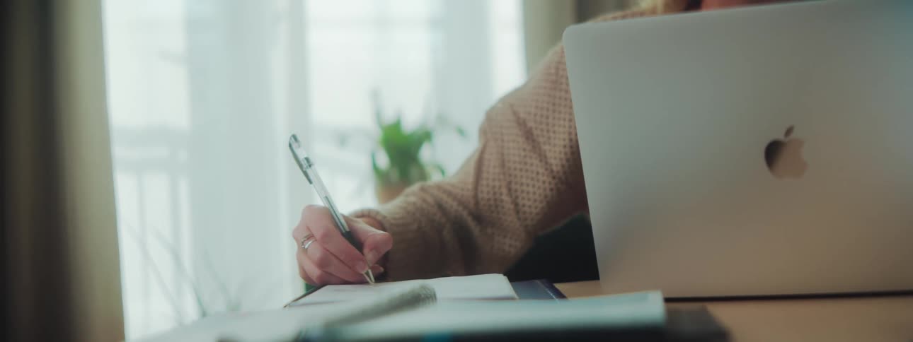 A woman sitting at a desk in front of a laptop is writing in a notebook