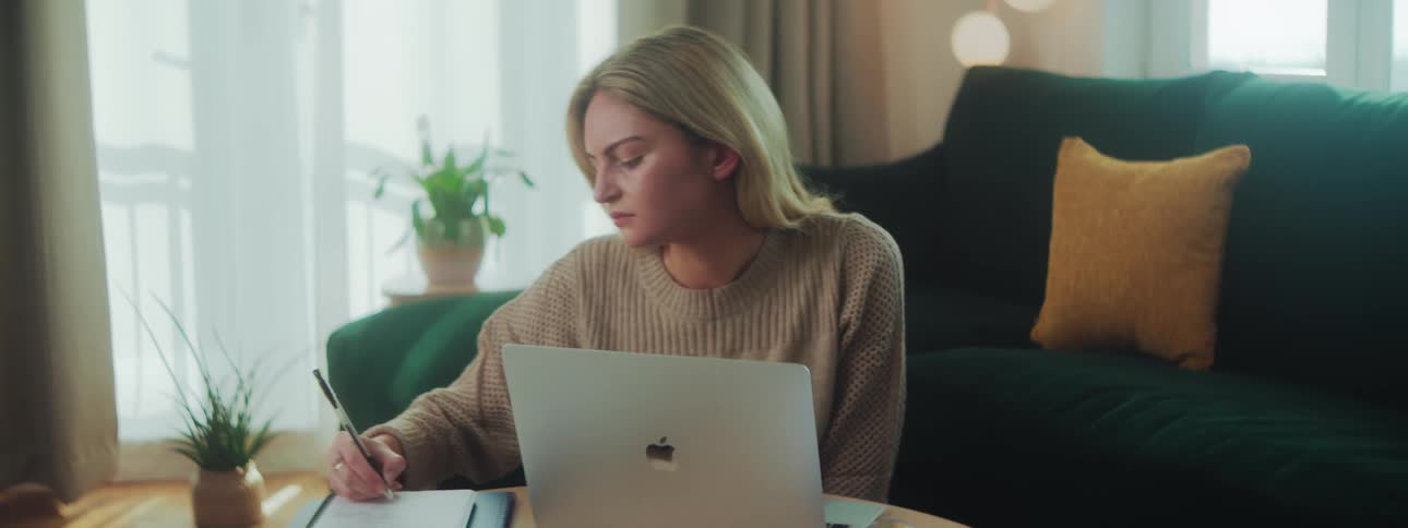 A woman is sitting on the floor in front of her couch working on a laptop and notebook on her coffee table