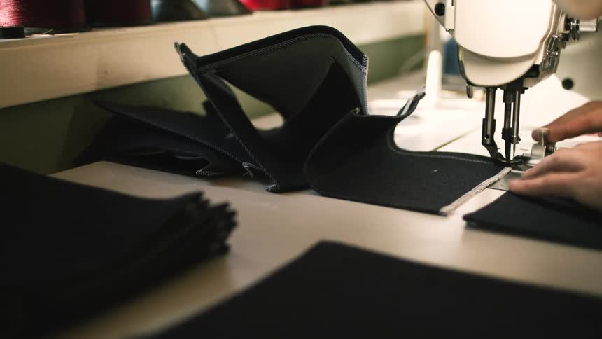 Someone operates a sewing machine stitching dark fabric on a worktable surrounded by thread spools and additional fabric