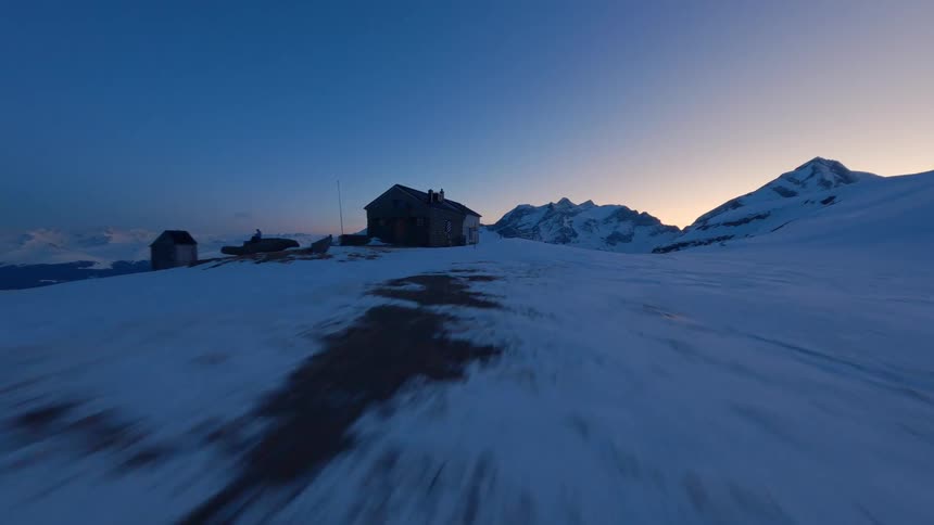 A smooth aerial shot glides over snowcovered mountain terrain at dusk eventually revealing a small isolated cabin nestled in the snowy landscape