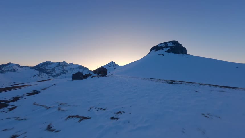 Snowy mountains and a house on top of a mountain with two people nearby at golden hour