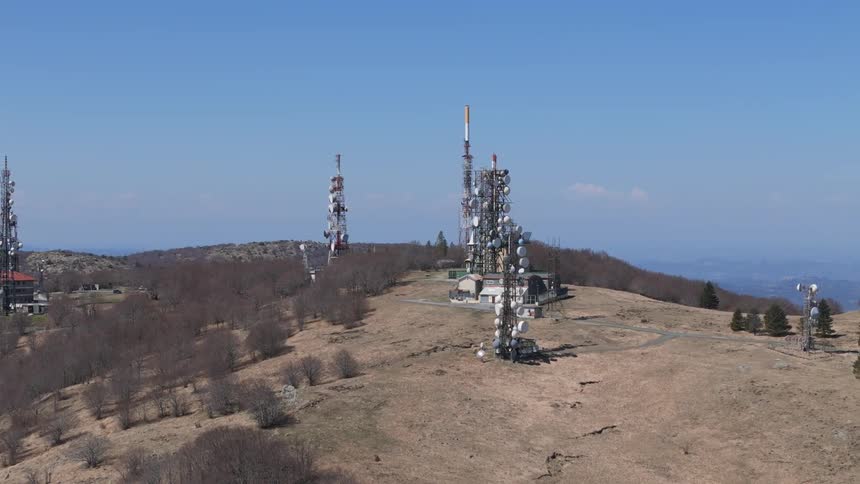 Aerial view orbits a communication station featuring multiple cell and broadcast towers equipped with antennas and microwave dishes situated on a sparse grassy hilltop under a clear blue sky