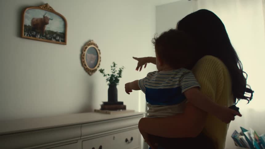 A mother holding her son in her arms is pointing at some paintings on the wall