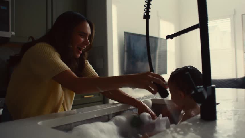 Happy baby splashing while bathing in kitchen sink as laughing mom sprays him with the faucet