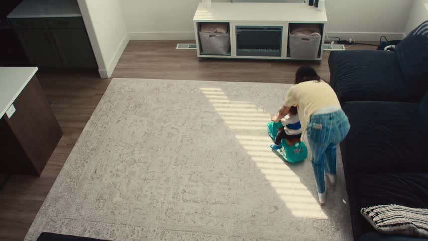 A mother plays by pushing her sons toy scooter on the living room rug