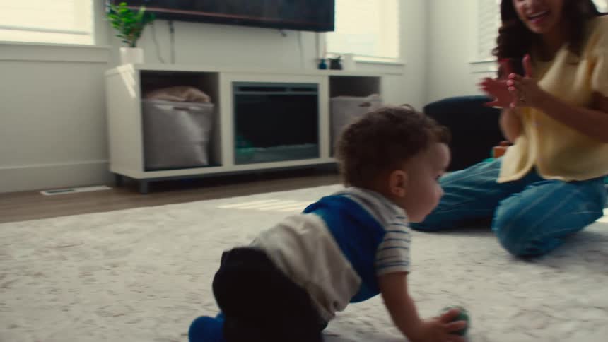 Baby picks up a ball and crawls to the right and drops it in front of a woman